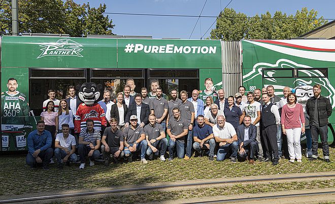 Im Vordergrund und mittig posiert eine große Gruppe von Männern und Frauen zusammen mit einem Panther-Maskottchen für ein Foto vor einer grünen und weißen Straßenbahn mit der Aufschrift "PANTHER" und "#PUREEMOTION".