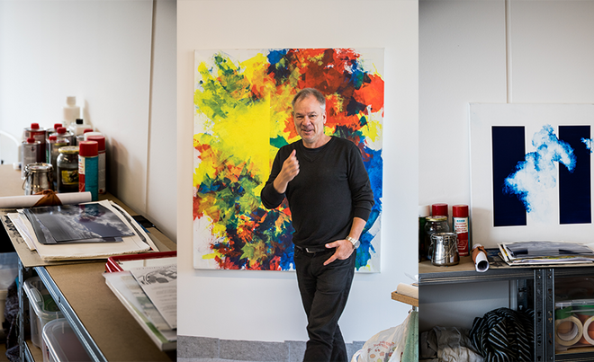 In einem Atelier steht ein Mann mittleren Alters in schwarzer Kleidung und macht mit dem Daumen ein „Daumen hoch“-Zeichen in die Kamera. Hinter ihm, an der Wand, hängt ein großes, abstraktes Gemälde, das von ihm selbst stammen könnte. Das Bild zeigt leuchtende Farbkleckse in Gelb, Rot, Blau und Weiß.