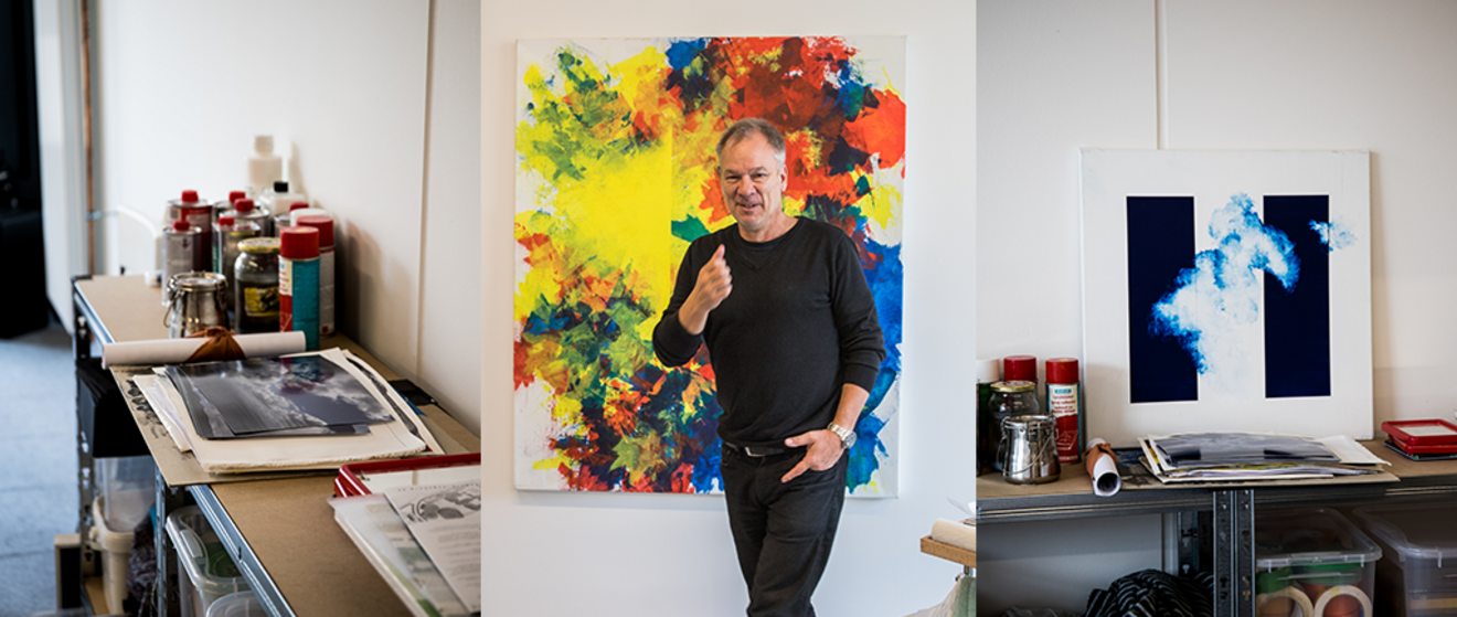 In einem Atelier steht ein Mann mittleren Alters in schwarzer Kleidung und macht mit dem Daumen ein „Daumen hoch“-Zeichen in die Kamera. Hinter ihm, an der Wand, hängt ein großes, abstraktes Gemälde, das von ihm selbst stammen könnte. Das Bild zeigt leuchtende Farbkleckse in Gelb, Rot, Blau und Weiß.