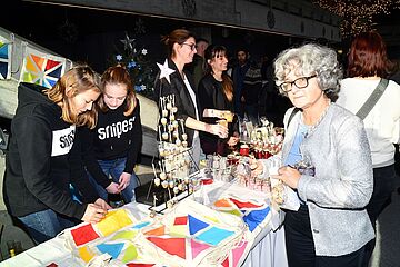 Eine festliche Szene, die einen Marktstand mit handgefertigten Artikeln auf einer Veranstaltung oder einem Adventsmarkt zeigt. Mehrere Personen interagieren an dem Stand, darunter drei junge Frauen und eine ältere Frau, die sich für die ausgestellten Waren interessiert. Ein Klick auf das Bild führt zu einer größeren Ansicht.