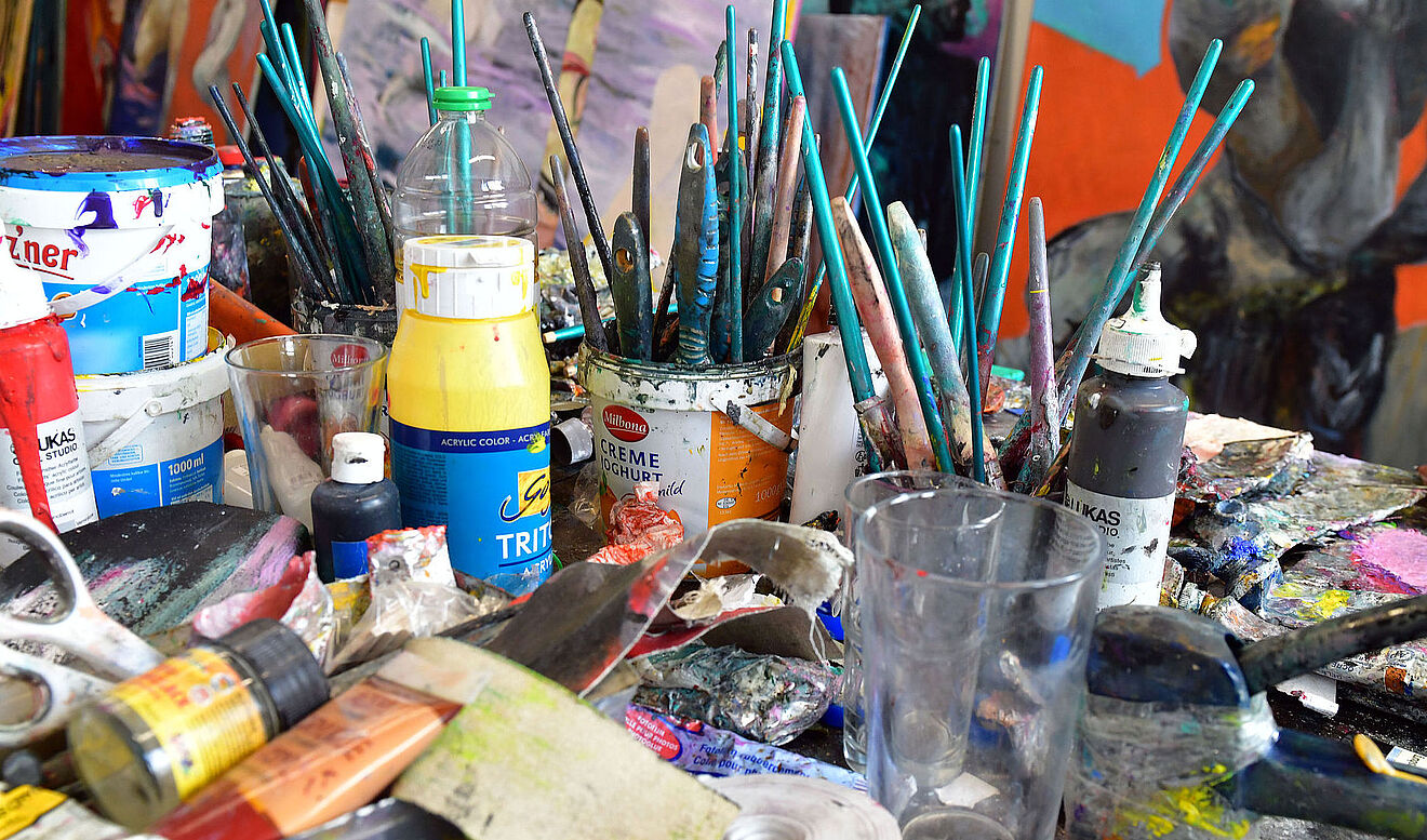 Voller Tisch mit Malutensillien. Unter anderem Farben, Scheren und Pinsel. Im Hintergrund sind Gemälde.