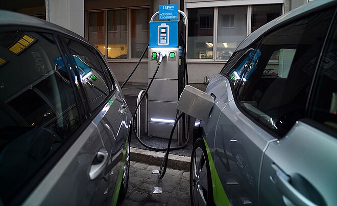 Zwei Carsharing-Autos der swa sind im Vordergrund. Eines davon ist an einer sich im Hintergrund befindenden eLadesäule angeschlossen.