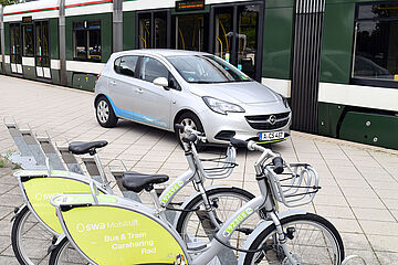Ein silbernes Auto und zwei silberne Fahrräder, alle mit swa-Logo versehen, stehen neben einer grün-weißen Straßenbahn an einem Bahnsteig. Ein Klick auf das Bild führt zu einer größeren Ansicht