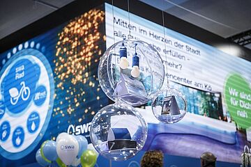 Drei hängende, transparente Kugeln mit innenliegenden Exponaten und kleinen Glühbirnen dominieren den Vordergrund eines Messestandes. Der große, beleuchtete Hintergrundbildschirm zeigt deutsche Slogans und Grafiken der SWA, umgeben von blauen und weißen Luftballons. Ein Klick auf das Bild führt zu einer größeren Ansicht.