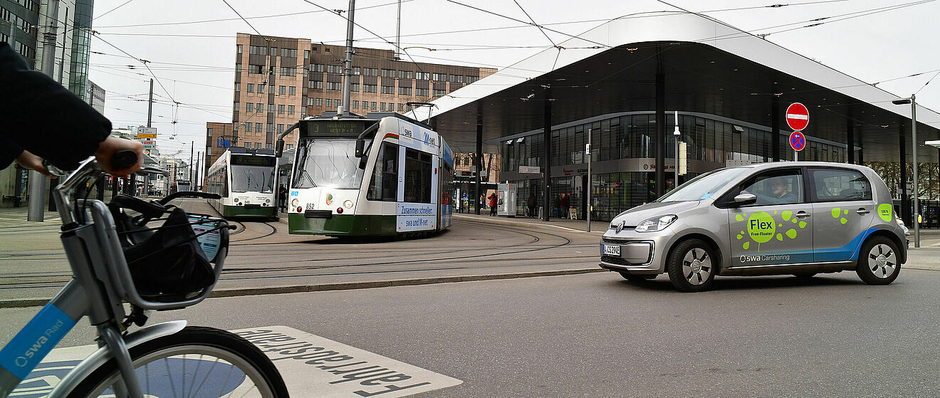 Foto vom Augsburger Königsplatz. Im Vordergrund zwei Trams, ein Carsharingauto und ein Leihrad.