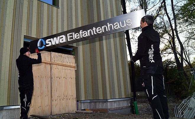 Montage eines Schilds am neuen swa Elefantenhaus im Zoo Augsburg.  Zwei Monteure in Arbeitskleidung, installieren das Schild mit der Aufschrift "swa Elefantenhaus".