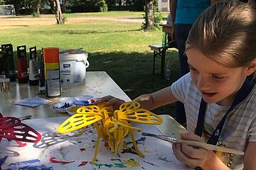 Ein junges Mädchen bemalt an einem Holztisch mit einem Pinsel eine große gelbe Wespe aus Holz oder Karton, umgeben von Bastelmaterialien. Ein Klick auf das Bild führt zu einer größeren Ansicht.