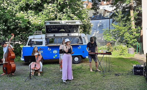 Eine Band gibt ein Hofkonzert, im Hintergrund steht der swa Bulli. 