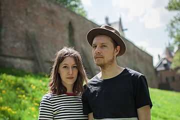 Isabel und Jakob stehen auf einer Wiese und schauen in die Kamera. Hinter ihnen befindet sich eine Mauer. - Ein Klick auf das Bild führt zur vergrößerten Fotoansicht.
