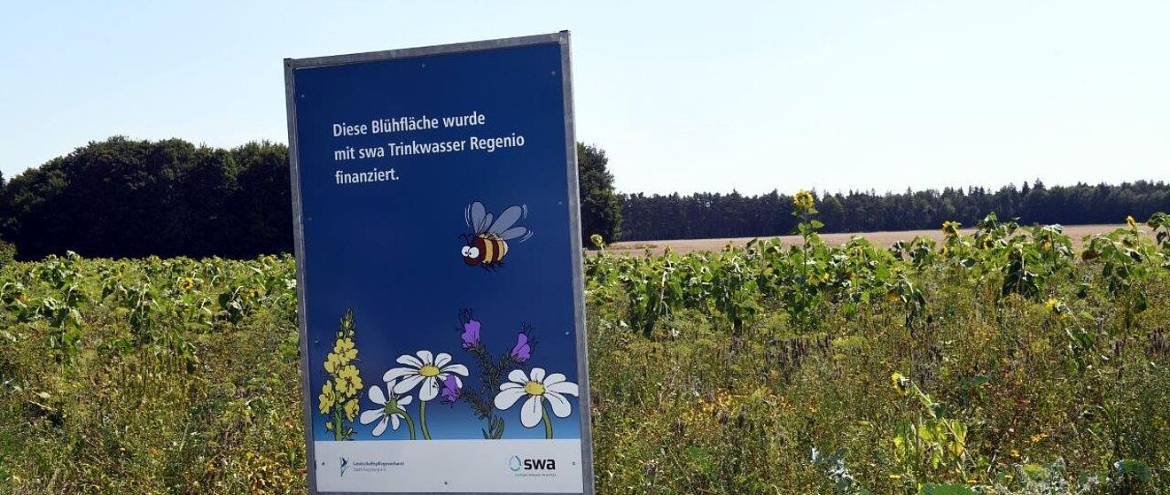 Ein Feld im Hintergrund, davor ein blaues Schild mit einer Darstellung einer Biene und Blumen. Auf dem Schild steht „Dieser Blühacker wurde mit Stadtwerke Trinkwasser Regenio finanziert.“
