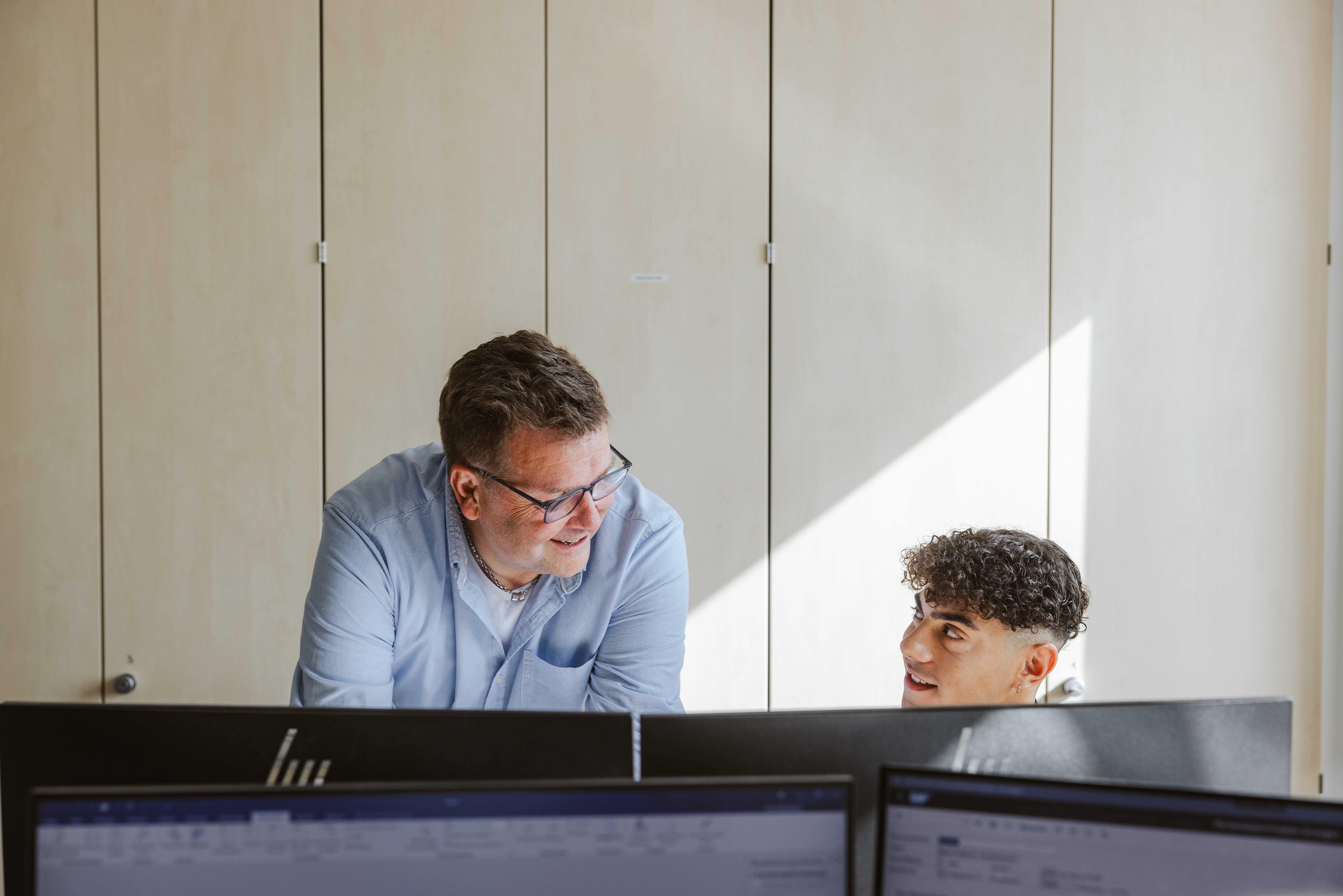 Ein Mann sitzt am Schreibtisch vor einem Monitor und unterhält sich mit einer stehenden Person neben ihm